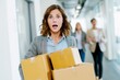 © id512 - Shocked young businesswoman struggling to carry multiple boxes in modern office hallway. Female employee drops package while coworkers walk behind, surprised expression on face.