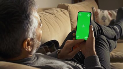 Man relaxes on couch using smartphone with green screen display.