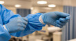 © MIQ - A healthcare professional carefully puts on blue disposable gloves over the sleeves of a protective gown in a medical setting, preparing for patient care.