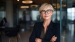 © Caspernik - Confident Caucasian female senior executive with stylish blonde hair and glasses posing with arms crossed in modern corporate office with blurred background of workspace and pendant lighting
