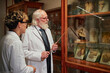 © Seventyfour - Senior Caucasian man with long beard and glasses explaining preserved specimens in glass cases to young adult man in laboratory coat inside museum setting
