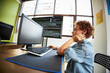 © DragonImages - Teenager Caucasian boy sitting at desk using desktop computer, holding neck with one hand, looking at monitor displaying programming code, modern office environment visible