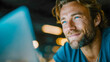 © Aliaksandra - Smiling man with blue eyes looking at a laptop, illuminated by warm bokeh lights