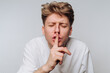 © bodnarphoto - Young man gestures for silence while wearing a white shirt against a neutral background during a quiet moment