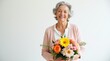 © Alex Pios - Elegant elderly woman with gray hair wearing soft pastel cardigan holds colorful birthday bouquet of mixed flowers including roses and daisies, genuine smile, bright natural lighting, clean white back