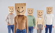 © Studio Romantic - Person with paper bag over head with happy emoticon mouth and eyes facial expression drawn on it. Casual young woman in smiling package bag standing in gray studio with group of people in background