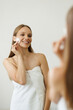 © F8  \ Suport Ukraine - Young beautiful woman cleaning her face skin with cotton pad in bathroom standing in towel, looking in the mirror