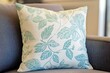© ALPADESIGN - A decorative throw pillow, featuring a subtle, muted teal leaf design on a light cream background, is displayed on a gray couch.
