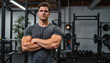© Elenba - Stern man with arms crossed in a gray shirt poses in a gym environment with weight lifting equipment and plants visible in the background, a motivational photo used for fitness concepts.