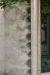 Modern vertical metal stand with multiple nozzles spraying fine water mist, located outdoors near green foliage, providing cooling or humidifying effect.