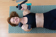 © Lana Pietukhova - A happy woman smiles and looks at the camera while lying on a mat, lifting two dumbbells, concept of fitness, health, and a happy lifestyle.