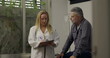 © Marco - Female doctor enters medical office to speak with male patient, beginning clinical conversation while holding clipboard, patient listens with serious expression
