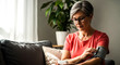 © @TrendStudio - An older woman with glasses and a pink shirt uses a blood pressure monitor on her arm She is seated on a sofa looking down at the device in a comfortable home setting