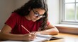 © Tetiana - Focused hispanic female teen studying by window with notebook and pencil