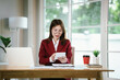 © Natthawadee - An Asian businesswoman in a suit works at her office desk with laptop and documents, analyzing finance, strategy, charts, and reports.