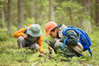 © Seventyfour - Two children exploring forest floor, one boy with blue backpack using magnifying glass to examine plants while another child in background observing nature closely