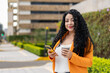 © Jordan - Happy Latin woman holding takeaway coffee and smartphone, city background