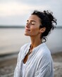 © Таня Андреева - Serene woman with freckles and curly hair smiling peacefully with eyes closed, enjoying warm sunlight by the ocean. Mindfulness and wellbeing concept.