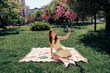 © deagreez - Lovely woman enjoying a sunny day in the park while taking a selfie on a blanket with colorful flowers around