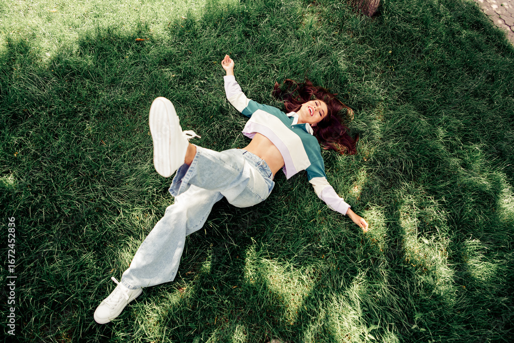 Young woman lying on grass outdoors in trendy casual outfit during spring, expressing joy and relaxation in natural light