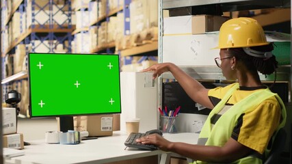 Female african american worker using pc with isolated mockup display, wearing hi vis vest and helmet in warehouse environment. E-commerce retail operations surrounded by big boxes. Camera B.