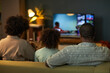 © DragonImages - Black family sitting on sofa watching television together, showing back view of woman, man, and child with curly hair focusing on screen in living room