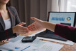 © MINAE - Two businesswomen shaking hands after reaching an agreement, with financial charts, calculator, and laptop on wooden table, celebrating successful partnership and deal