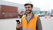 © Denis Yevtekhov - A smiling worker in safety gear using a smartphone at an industrial site, logistics worker, industrial operations, digital management, global trade, freight hub