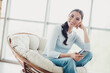 © deagreez - Beautiful woman seated comfortably with a smartphone smiles in a cozy chair against a bright indoor backdrop