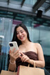 © PaeGAG - Young woman using mobile phone while holding shopping bags in mall