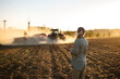 © encierro - Farmer with smartphone monitoring smart farming cultivation while tractor and seeder prepare soil during agricultural work on field
