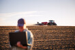 © encierro - Smart farming and modern agriculture with farmer using laptop while tractor with seeder sowing crop seeds on field. Agronomist with computer analyzing farm data