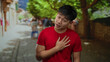 © Krakenimages.com - Young man in red shirt stands on city street expressing emotion with hand on chest, surrounded by blurred people and trees under bright outdoor lighting