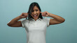 © Krakenimages.com - Woman covering ears standing against blue background displaying frustration and discomfort, wearing white shirt, young latin female with expression of annoyance.