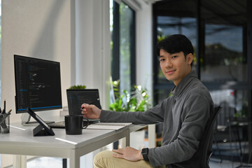 Portrait of a Young programmer smiling at the camera while working on code in a bright, modern office environment