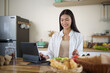 © matinee - Smiling young woman using a digital tablet while having healthy breakfast with salad and croissants in kitchen.