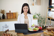 © matinee - Smiling young woman using a digital tablet while having healthy breakfast with salad and croissants in kitchen.
