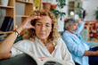 © we.bond.creations - Mature woman with curly hair reading a book while touching her forehead and her husband is pensive on the sofa in the background