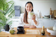 © matinee - Happy Asian woman enjoying homemade matcha green tea latte in a modern kitchen.