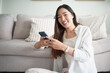 © matinee - Happy young asian woman sitting on floor and using smartphone at home.