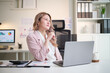 © wattana - Professional businesswoman looking thoughtful while working on a laptop with financial reports on desk.