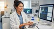 © patricia - A smiling woman in a lab coat sits at a desk typing on a keyboard while looking at a computer screen displaying graphs