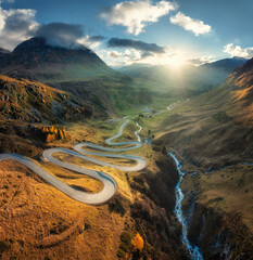 Wall Mural - Aerial view of winding mountain road in the Swiss Alps, Switzerland, with sharp curves, golden slopes, valley and dramatic sun rays breaking through clouds over alpine peaks. Top drone view of road