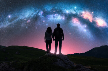 Wall Mural - Couple of hikers holding hands standing on a rocky outcrop in mountains, under pink night starry sky with glowing Milky Way. Landscape with silhouettes of man and woman, bright stars and milky way