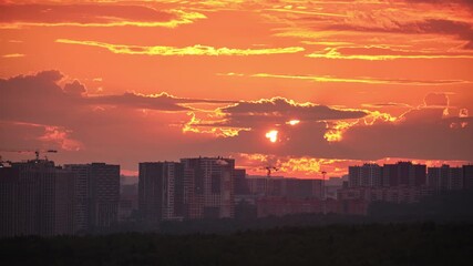 Wall Mural - A vivid orange sunset casts dramatic light over a city skyline with silhouetted high-rise buildings and cranes against glowing clouds, time lapse.