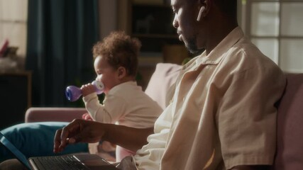 Cinematic side view shot of young African American father caring for baby while working from home and using laptop, cozy domestic scene lit by sunlight, copy space