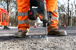 © Iryna - Construction worker in orange clothes and safety boots using cordless drill to break asphalt on road project in rural area