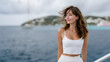 © hasara - Relaxed young woman in a white crop top and skirt smiling peacefully while standing on a boat, with a scenic view of the ocean, sailboats, and coastal cliffs in the background