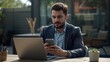 © Ajigus - A man in a suit sits outdoors, looking intently at his mobile phone while working on his laptop.