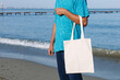 © makanna - Man with black, tan skin holding in hand white eco cotton textile bag. Blue water of sea or ocean coast background. Seaside, sunny day. Blank mock up tote bag, eco concept, place for text design logo